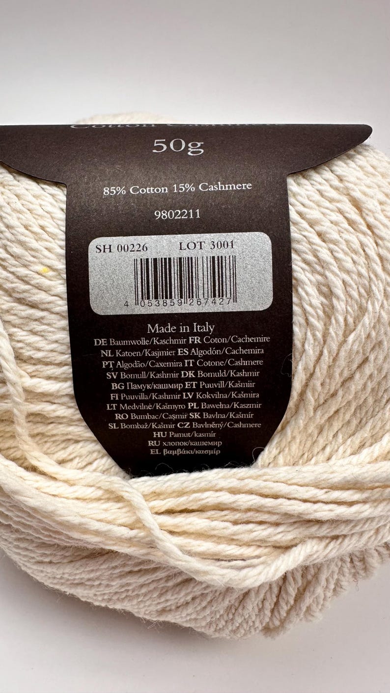 May include: A ball of off-white yarn with a dark brown paper label. The label states 50g, 85% Cotton 15% Cashmere, and Made in Italy. It includes a barcode, lot number 3001, and product code 9802211. The yarn has a thick, twisted texture.