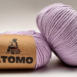 May include: Two skeins of lavender-colored yarn. One skein has a brown label with the word "ATOMO" and an illustration of a person. The yarn is tightly wound, showing the texture of the fibers. Ideal for knitting and crochet projects.