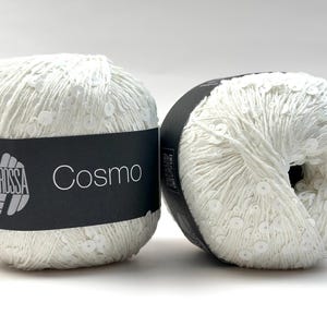 May include: Two white yarn balls with a sequin-like texture. The yarn is wrapped around a black label with the word "Cosmo" and the Lana Grossa logo. The yarn balls are on a white background.