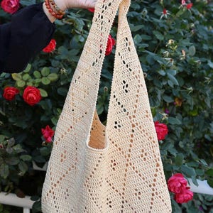 May include: A cream-colored crochet hobo bag featuring an openwork diamond pattern throughout the body and a single integrated shoulder strap. The bag is constructed from yarn with a visible textured stitch pattern and a soft, flexible structure.
