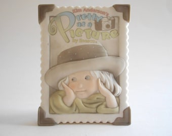 Może przedstawiać: Porcelanowa figurka dziewczynki z blond włosami, w brązowym kapeluszu i żółtej sukience. Figurka jest częścią kolekcji "Pretty as a Picture" Kim Anderson.