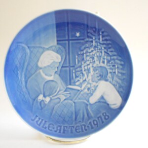May include: A blue and white ceramic Christmas plate with a scene of a woman reading to a child in front of a Christmas tree. The plate has the text "Juleaften 1978" on the bottom.