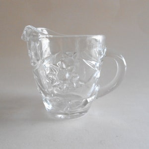 May include: A clear glass creamer with a floral pattern. The creamer has a handle and a spout.