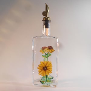 May include: Clear glass bottle with a sunflower design. The bottle has a metal spout and a cork stopper. The sunflower is painted in yellow, brown, and green. The bottle is rectangular and stands upright.