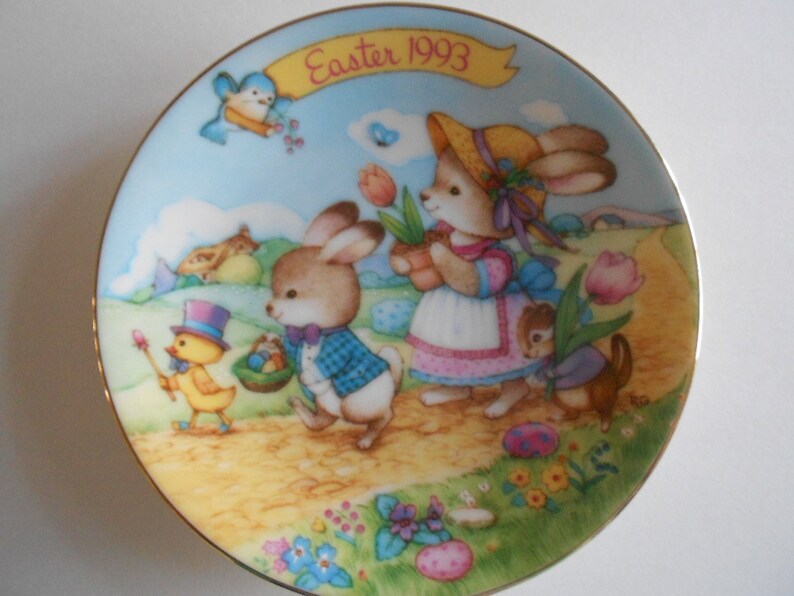 May include: A porcelain plate with a gold rim features a colorful illustration of bunnies, a chick, and a squirrel. The plate has the text "Easter 1993" on a banner.
