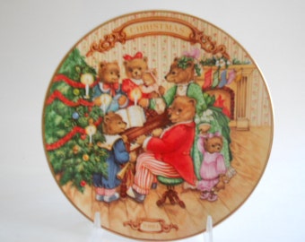 Może przedstawiać: Ceramiczny talerz świąteczny z przedstawieniem rodziny niedźwiedzi śpiewających kolędy wokół choinki. Talerz przedstawia zielone drzewko z czerwonymi i złotymi ozdobami, kominek z pończochami oraz rodzinę niedźwiedzi ubranych w świąteczne stroje. Tekst "Christmas" jest napisany złotymi literami na górze talerza.