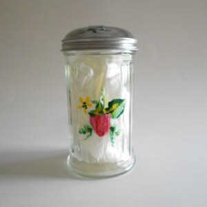 May include: A clear glass sugar dispenser with a silver lid. The dispenser has a painted design of a strawberry, leaves, and yellow flowers.