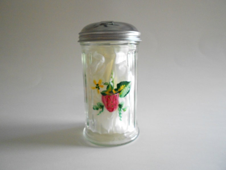 May include: A clear glass sugar dispenser with a silver lid. The dispenser has a painted design of a strawberry, leaves, and yellow flowers.