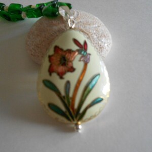 Pu&ograve; includere: Un ciondolo in smalto bianco con un motivo floreale verde, blu e arancione. Il ciondolo &egrave; attaccato a una collana di perline verdi.