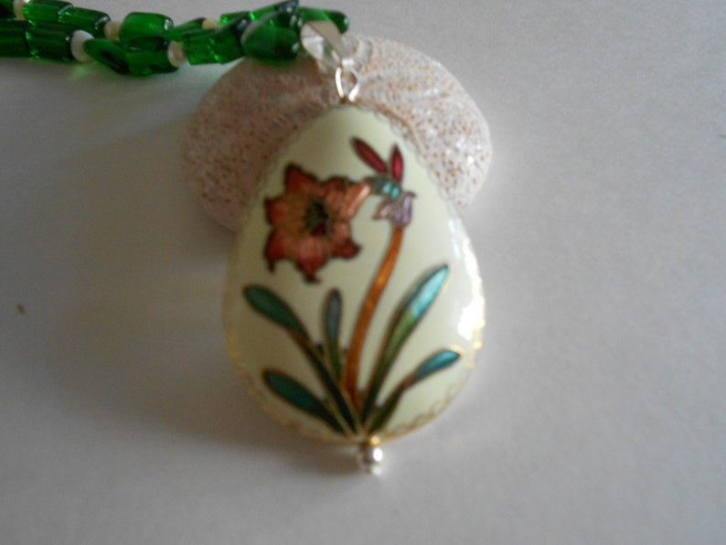 Pu&ograve; includere: Un ciondolo in smalto bianco con un motivo floreale verde, blu e arancione. Il ciondolo &egrave; attaccato a una collana di perline verdi.