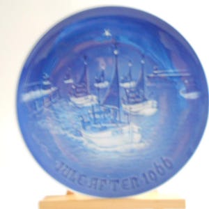 May include: Blue and white ceramic plate with a raised design of ships sailing on a blue sea. The plate has the text "Julgratter 1968" on the bottom.