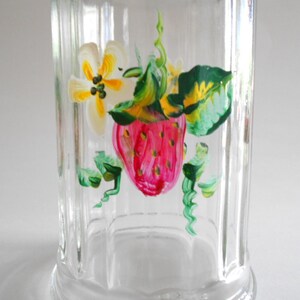May include: A clear glass jar with a hand-painted design of a strawberry, leaves, and a yellow flower. The jar has a ribbed texture.