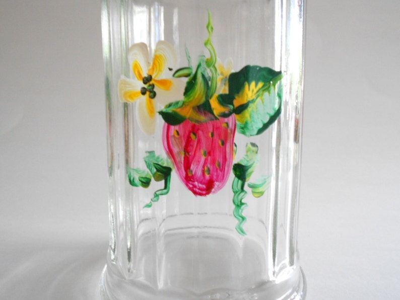 May include: A clear glass jar with a hand-painted design of a strawberry, leaves, and a yellow flower. The jar has a ribbed texture.