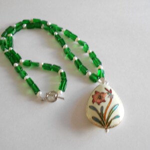 Pu&ograve; includere: Una collana di perline di vetro verde e bianco con un ciondolo che presenta un motivo floreale in tonalit&agrave; di rosa, verde e oro.