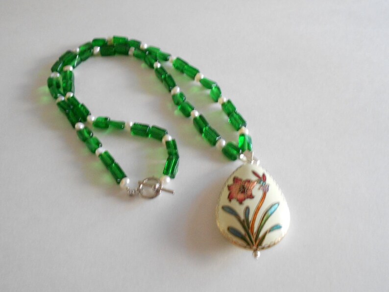 Pu&ograve; includere: Una collana di perline di vetro verde e bianco con un ciondolo che presenta un motivo floreale in tonalit&agrave; di rosa, verde e oro.