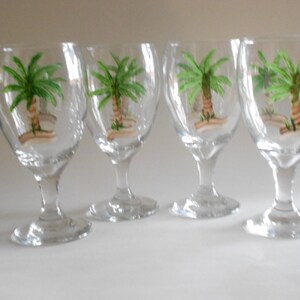 May include: Set of four clear glass goblets with painted palm tree designs. Each goblet has a round bowl and a tall, slender stem.
