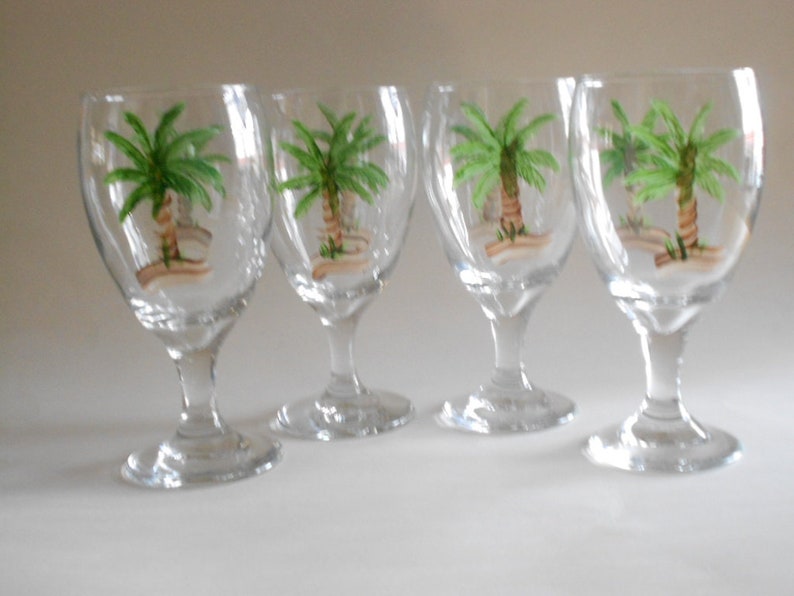 May include: Set of four clear glass goblets with painted palm tree designs. Each goblet has a round bowl and a tall, slender stem.