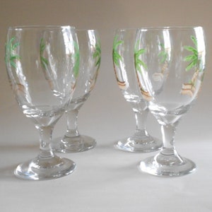 May include: Four clear glass wine glasses with painted palm trees on the side. The glasses have a round bowl and a long stem.