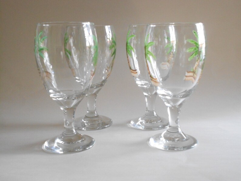 May include: Four clear glass wine glasses with painted palm trees on the side. The glasses have a round bowl and a long stem.