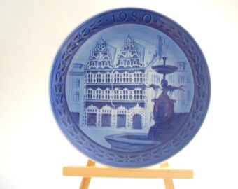 Może przedstawiać: Okrągły talerz kolekcjonerski z niebiesko-białej porcelany z ilustracją historycznego budynku i fontanny. Rok 1980 jest wytłoczony u góry. Talerz posiada ozdobny, ażurowy brzeg z powtarzającym się motywem liści.