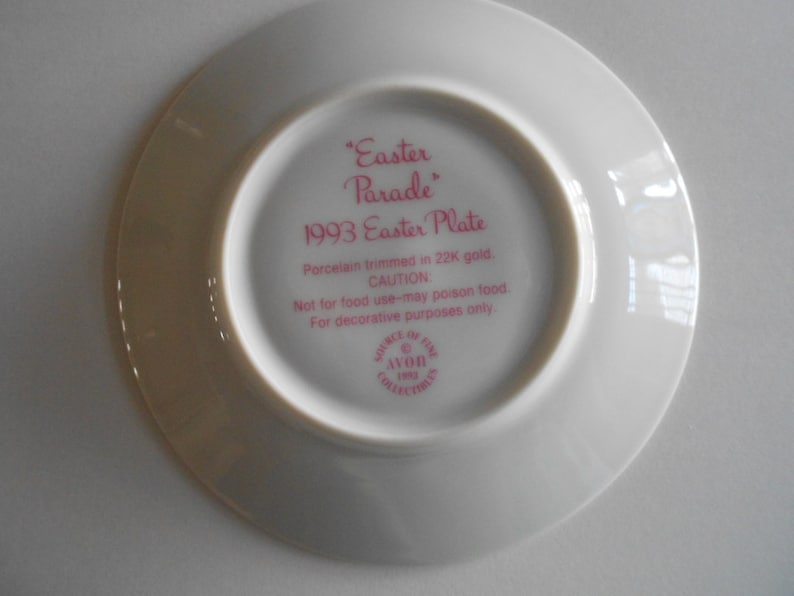 May include: White porcelain plate with a pink rim and pink text that reads "Easter Parade 1993 Easter Plate". The text continues with "Porcelain trimmed in 22K gold. CAUTION Not for food use - may poison food. For decorative purposes only." The plate also has a small pink circle with the text "Avon Collectors 1993" in white.