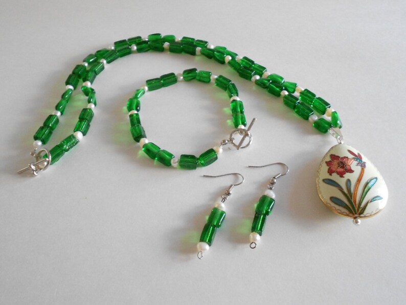 Pu&ograve; includere: Una collana di perline verdi e bianche con un ciondolo con un motivo floreale. Sono inclusi anche orecchini abbinati in perline verdi e bianche.