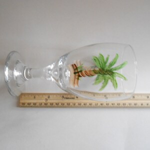 May include: A clear glass wine glass with a hand-painted palm tree design. The tree is painted in shades of green, brown, and yellow.