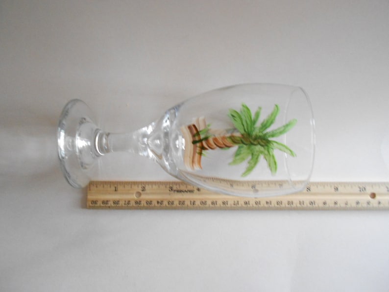 May include: A clear glass wine glass with a hand-painted palm tree design. The tree is painted in shades of green, brown, and yellow.