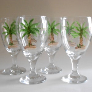 May include: Set of four clear glass wine glasses with hand-painted palm tree designs. Each glass features a green palm tree with a brown trunk and green leaves.