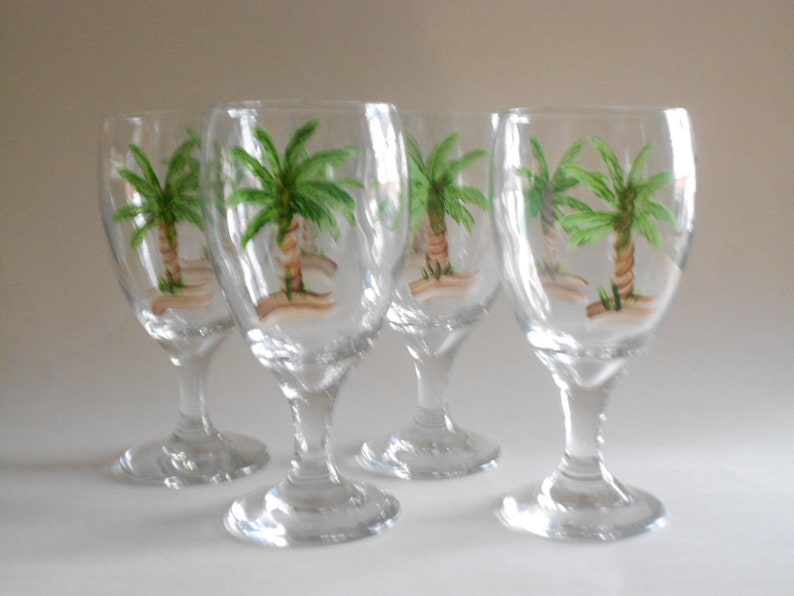 May include: Set of four clear glass wine glasses with hand-painted palm tree designs. Each glass features a green palm tree with a brown trunk and green leaves.