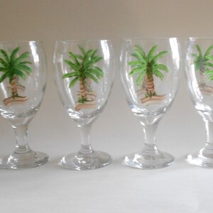 May include: Set of four clear glass wine glasses with hand-painted palm tree designs. Each glass features a green palm tree with a brown trunk and a white banner.