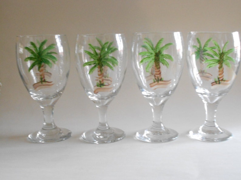 May include: Set of four clear glass wine glasses with hand-painted palm tree designs. Each glass features a green palm tree with a brown trunk and a white banner.