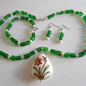 Pu&ograve; includere: Un set di collana e braccialetto con perline verdi e bianche con un ciondolo che presenta un motivo floreale. Il ciondolo &egrave; di forma ovale e ha uno sfondo bianco con un fiore rosso e giallo e foglie verdi.