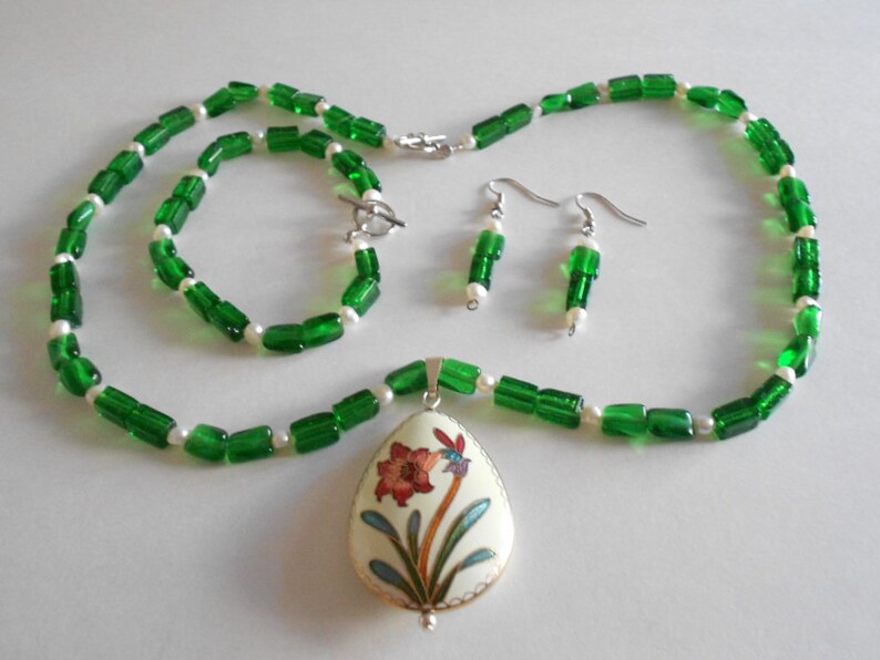 Pu&ograve; includere: Un set di collana e braccialetto con perline verdi e bianche con un ciondolo che presenta un motivo floreale. Il ciondolo &egrave; di forma ovale e ha uno sfondo bianco con un fiore rosso e giallo e foglie verdi.