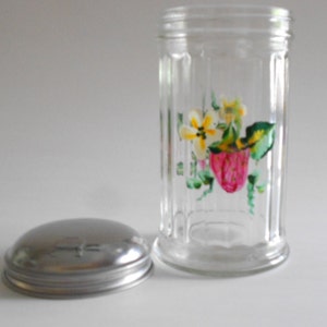 May include: A clear glass jar with a metal lid. The jar has a floral design painted on the side, featuring pink strawberries, yellow flowers, and green leaves.