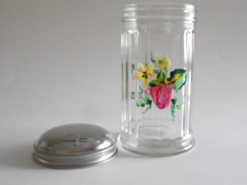 May include: A clear glass jar with a metal lid. The jar has a floral design painted on the side, featuring pink strawberries, yellow flowers, and green leaves.