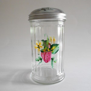 May include: A clear glass sugar dispenser with a silver lid. The dispenser has a hand-painted design of a strawberry and flowers in shades of pink, yellow, and green.