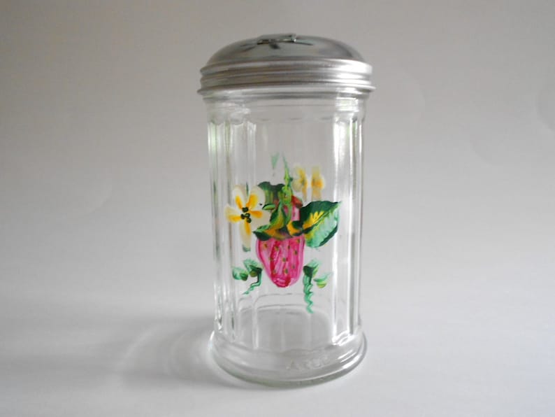 May include: A clear glass sugar dispenser with a silver lid. The dispenser has a hand-painted design of a strawberry and flowers in shades of pink, yellow, and green.