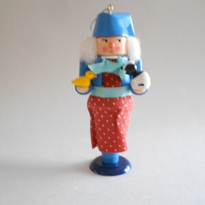 May include: A wooden nutcracker ornament with a blue hat, white fur trim, and a red and white polka dot dress. The nutcracker is holding a yellow bird and a black and white dog.