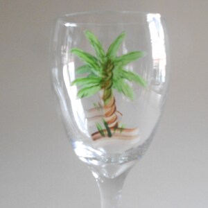 May include: A clear glass wine glass with a hand-painted palm tree design. The palm tree is green with a brown trunk and is painted on the front of the glass.