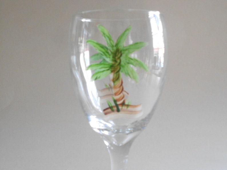 May include: A clear glass wine glass with a hand-painted palm tree design. The palm tree is green with a brown trunk and is painted on the front of the glass.