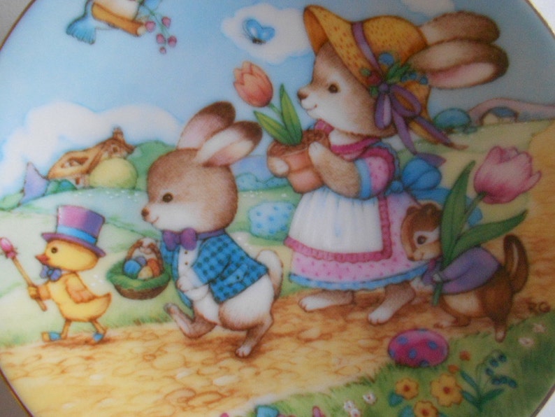 May include: A pastel-colored ceramic plate with a whimsical Easter scene. A yellow chick with a top hat holds a wand, while two bunnies, one wearing a straw hat and the other a blue plaid jacket, walk along a path. A brown squirrel carries a pink tulip.