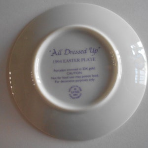 Pode incluir: Um prato de porcelana branco com uma inscri&ccedil;&atilde;o roxa que diz "All Dressed Up" 1994 Easter Plate. O prato &eacute; guarnecido com ouro de 22 quilates e &eacute; apenas para fins decorativos.