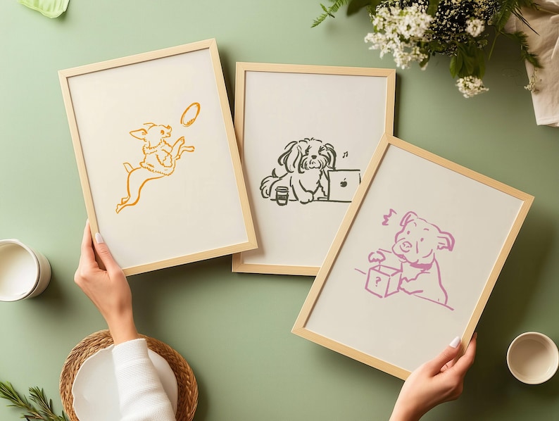 May include: Three light wood framed prints featuring minimalist line art illustrations of dogs. One print shows a yellow dog jumping for a disc, another a black dog at a laptop, and the third a pink dog interacting with a box marked with a question mark.