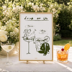 Może przedstawiać: Oprawiony znak z napisem "Drink on Us" i ilustracjami martini i whisky sour. Kieliszek do martini z oliwkami i koktajl whisky sour z wiśnią i plasterkiem pomarańczy znajdują się na stole.