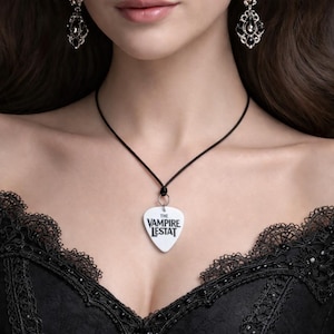 May include: A white guitar pick pendant with the text "THE VAMPIRE LESTAT" in black lettering hangs from a black cord necklace. The image shows a black lace top and ornate black earrings.