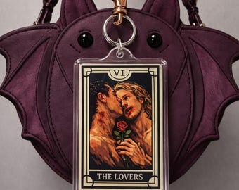 May include: A purple bat-shaped handbag with a gold chain strap and a keychain featuring a tarot card. The tarot card depicts two figures kissing, with the words "THE LOVERS" at the bottom. The bag has bat wings and small black eyes.
