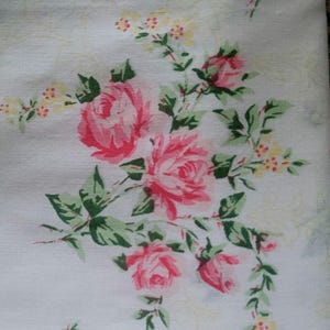 Könnte beinhalten: Ein weißer Stoff mit einem Blumenmuster aus rosa Rosen, grünen Blättern und kleinen gelben Blüten. Das Design ist in Gruppen angeordnet, wobei Stängel und Blätter nach außen ragen und ein sich wiederholendes botanisches Motiv bilden.