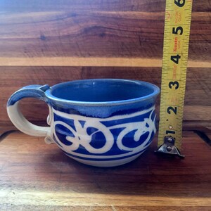 May include: A handmade ceramic mug with a blue interior and a white and blue decorative pattern. The mug has a handle and measures approximately 3 inches in diameter.