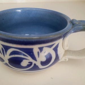 May include: A ceramic mug with a wide, rounded bowl shape and a sturdy handle. The interior and upper rim are glazed in a soft periwinkle blue, while the exterior features a deep cobalt blue band decorated with a white, hand-painted floral vine pattern.